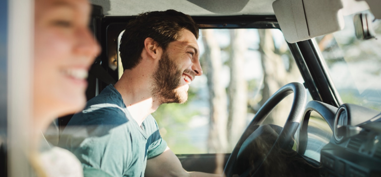 A smiling man driving a vehicle with a smiling woman sitting in the passenger seat as they both look forward. 