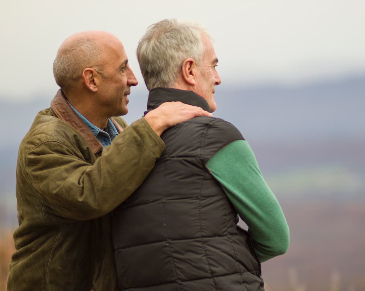 Two men embrace and stare into the distance together in a nature setting.