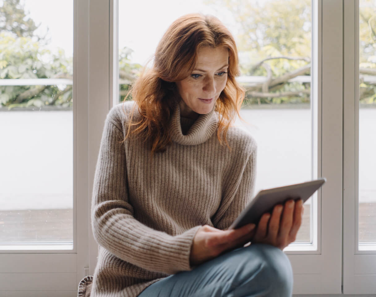 A woman with red hair and a beige sweater using a tablet.