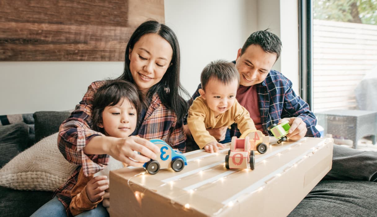 A family of four plays with toy cars in a living room setting.