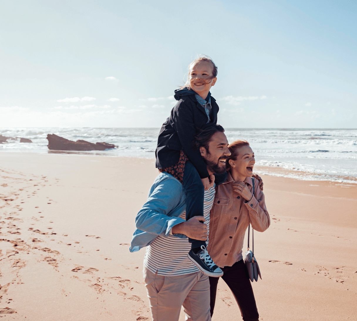 A family of three walking on the beach with blue skies, the ocean, and footprints in the sand. 
