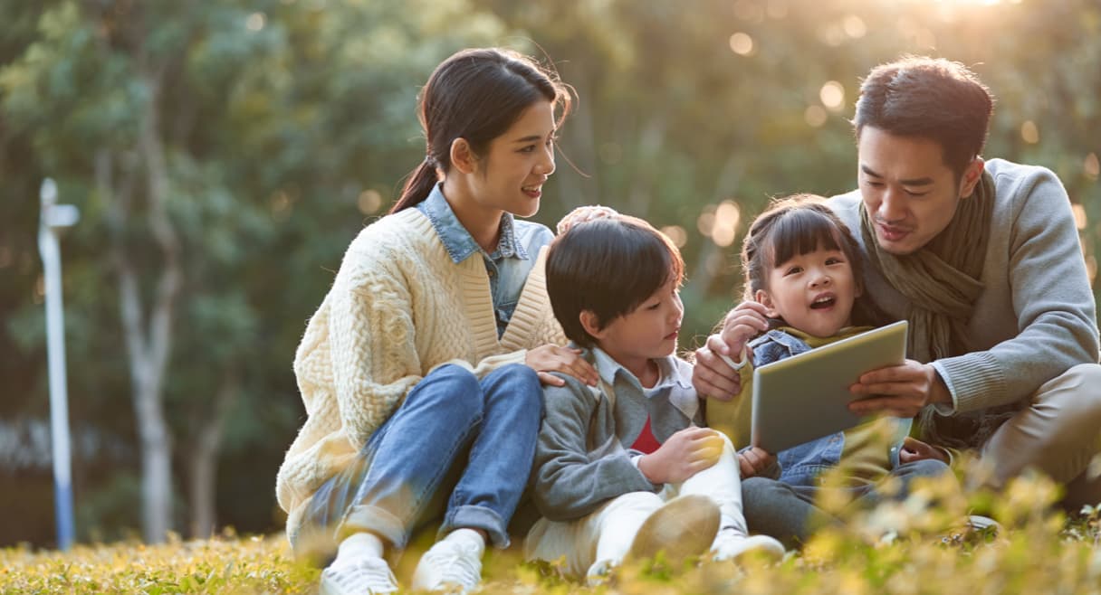 A family sitting outside together and sitting in a park with trees and yellow flowers looking at an iPad. 